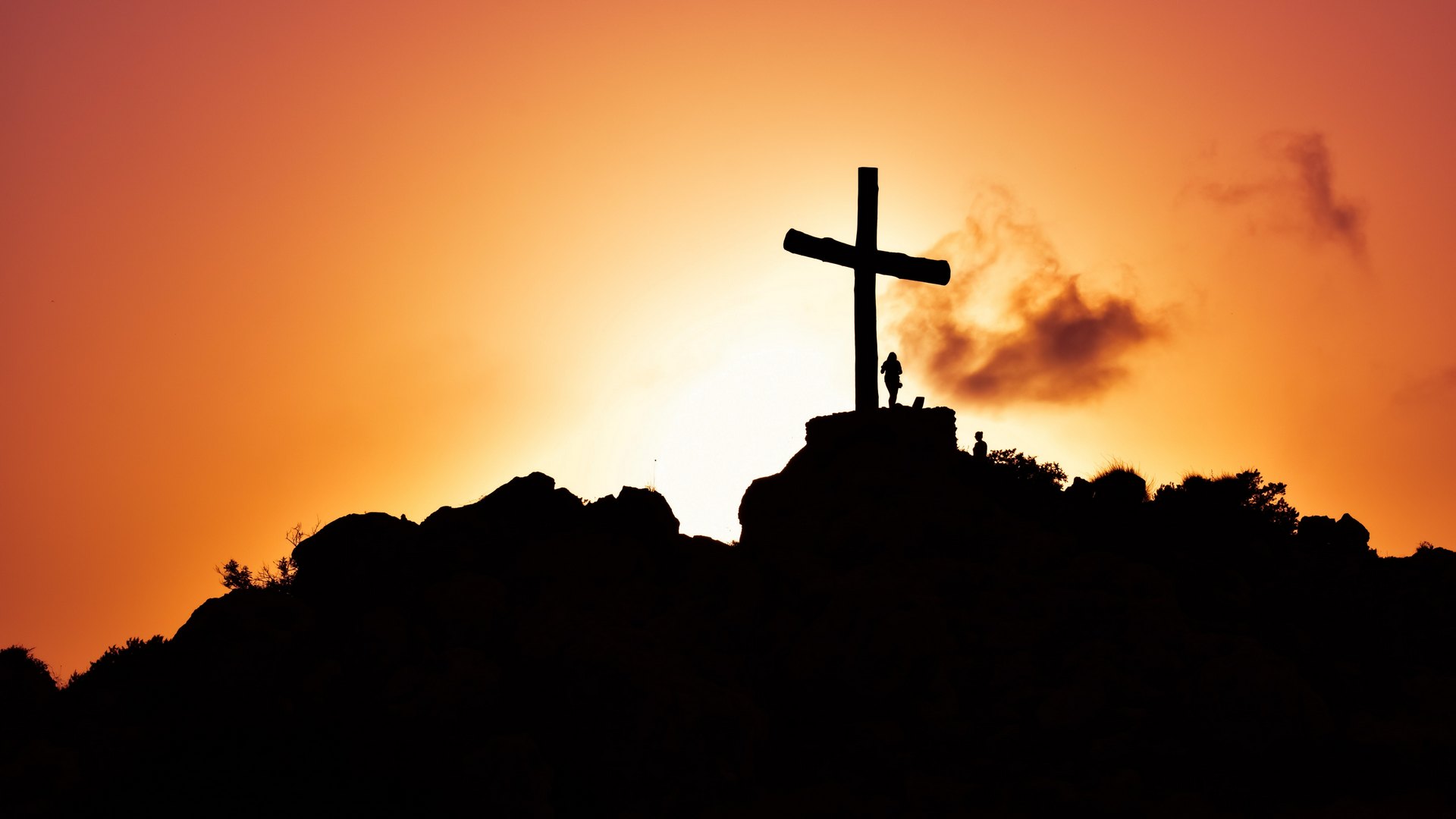 Silhouette photo of a mountain cross and a man standing underneath the cross with an orange glowing background